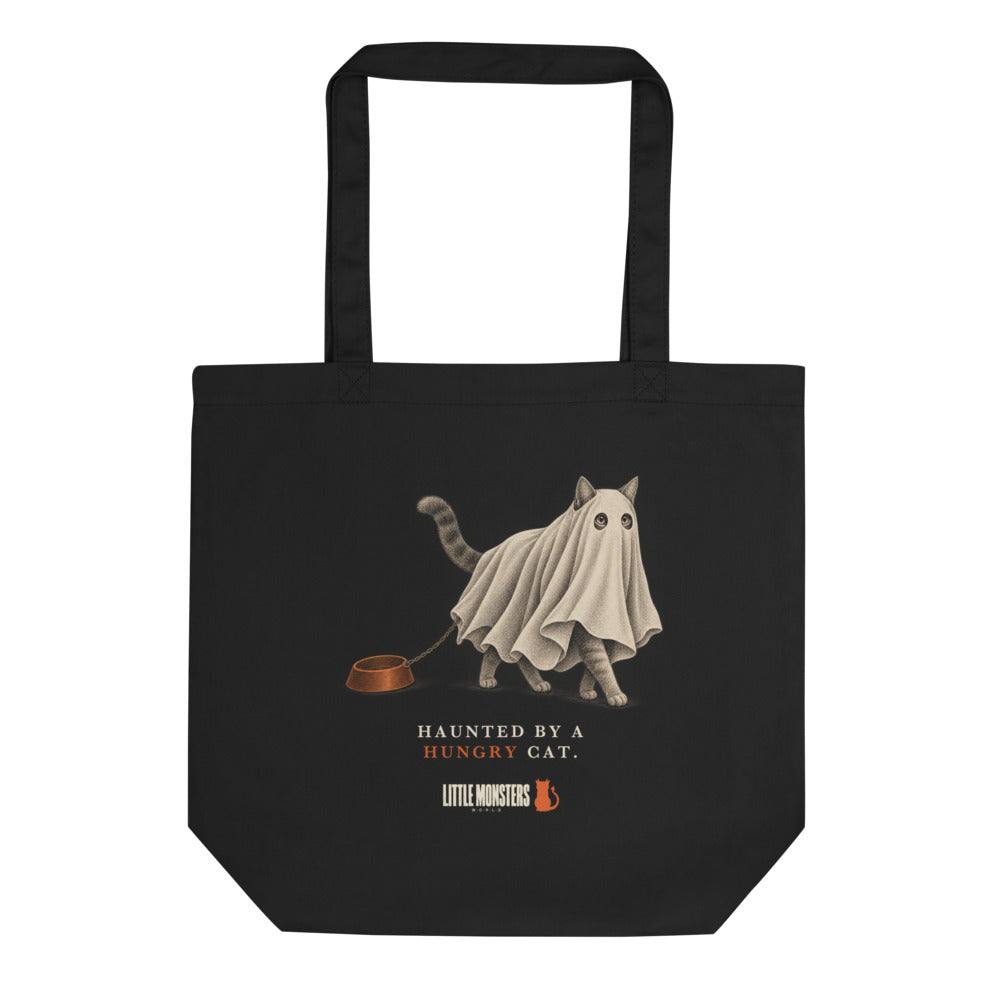 Black tote bag with a cat in a ghost sheet dragging its food bowl and the words Haunted by a hungry cat