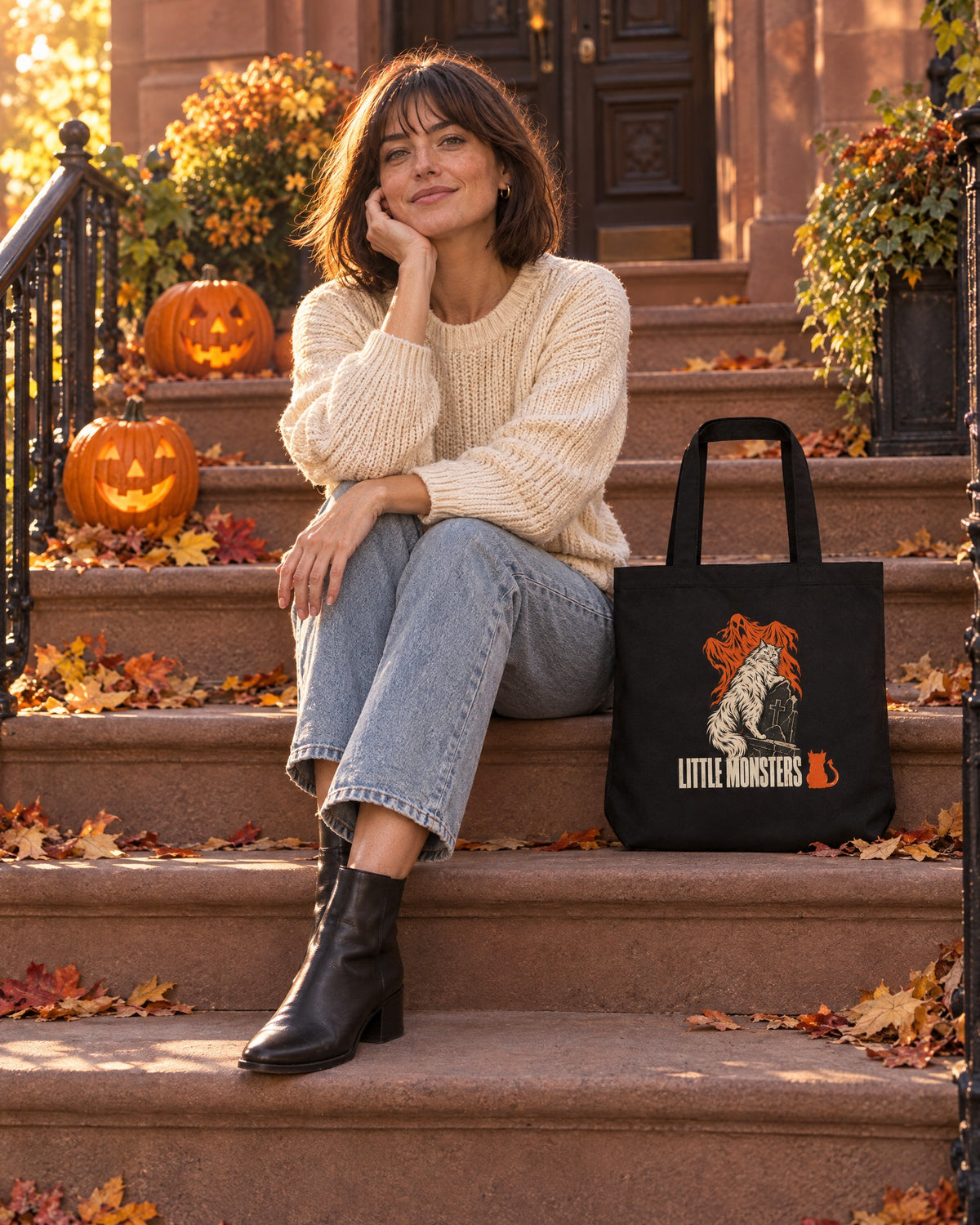 Woman in a cream sweater on a brownstone stoop with jack-o'-lanterns, the Little Monsters Halloween 2026 graveyard cat tote beside her