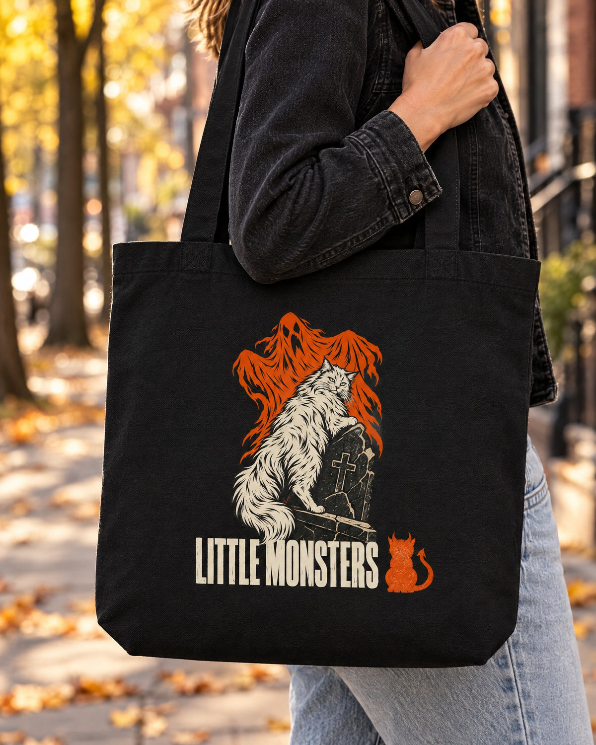 Close-up of the Halloween 2026 tote on the shoulder: a white long-haired cat on a gravestone with orange ghosts and the Little Monsters logo