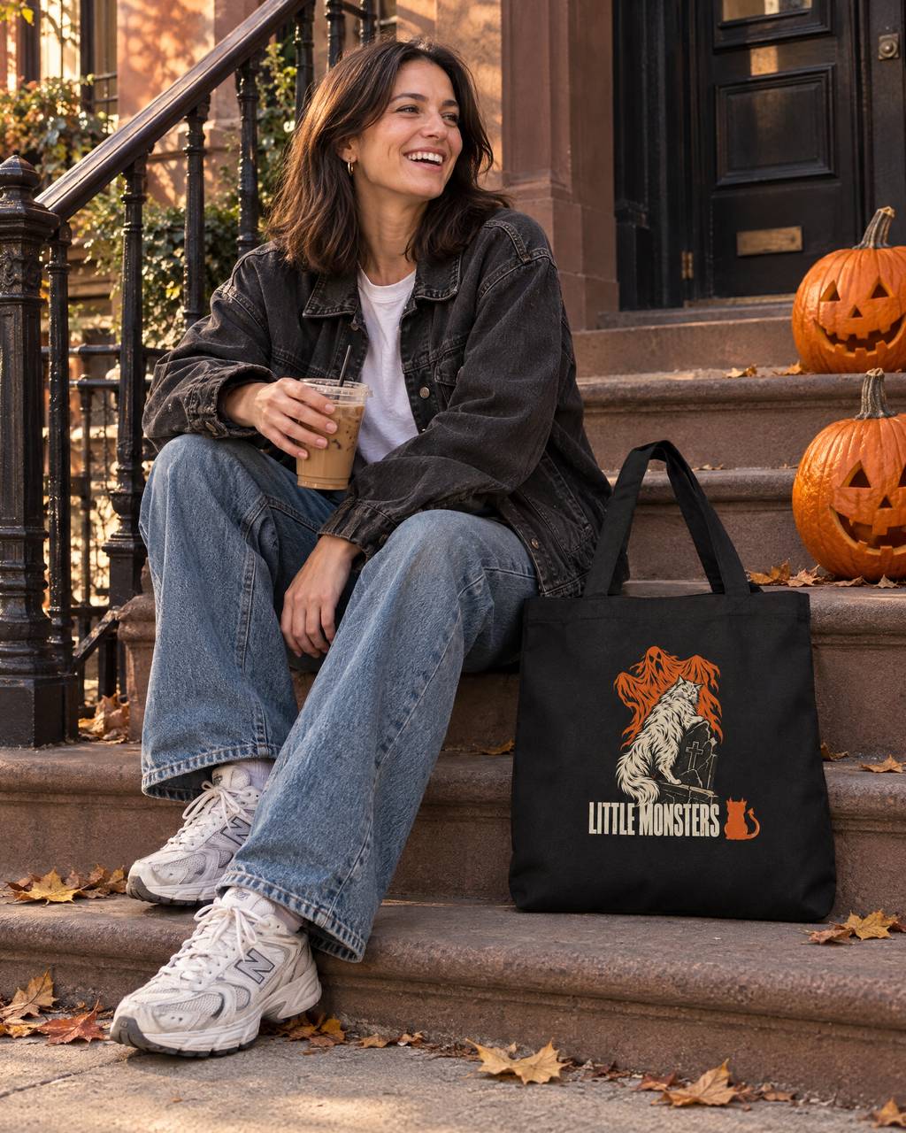 Woman on a brownstone stoop with pumpkins and the Little Monsters Halloween 2026 graveyard cat tote bag