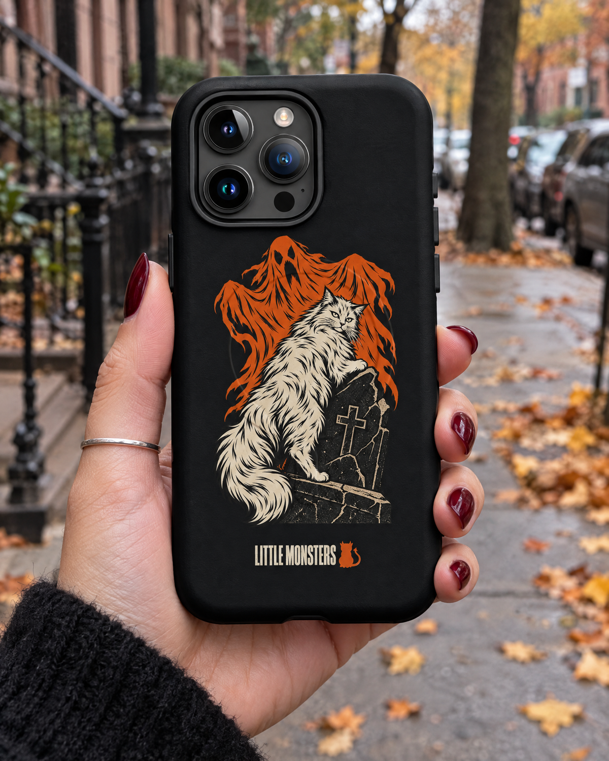 Close-up of a hand holding the Little Monsters Halloween 2026 graveyard cat MagSafe iPhone case