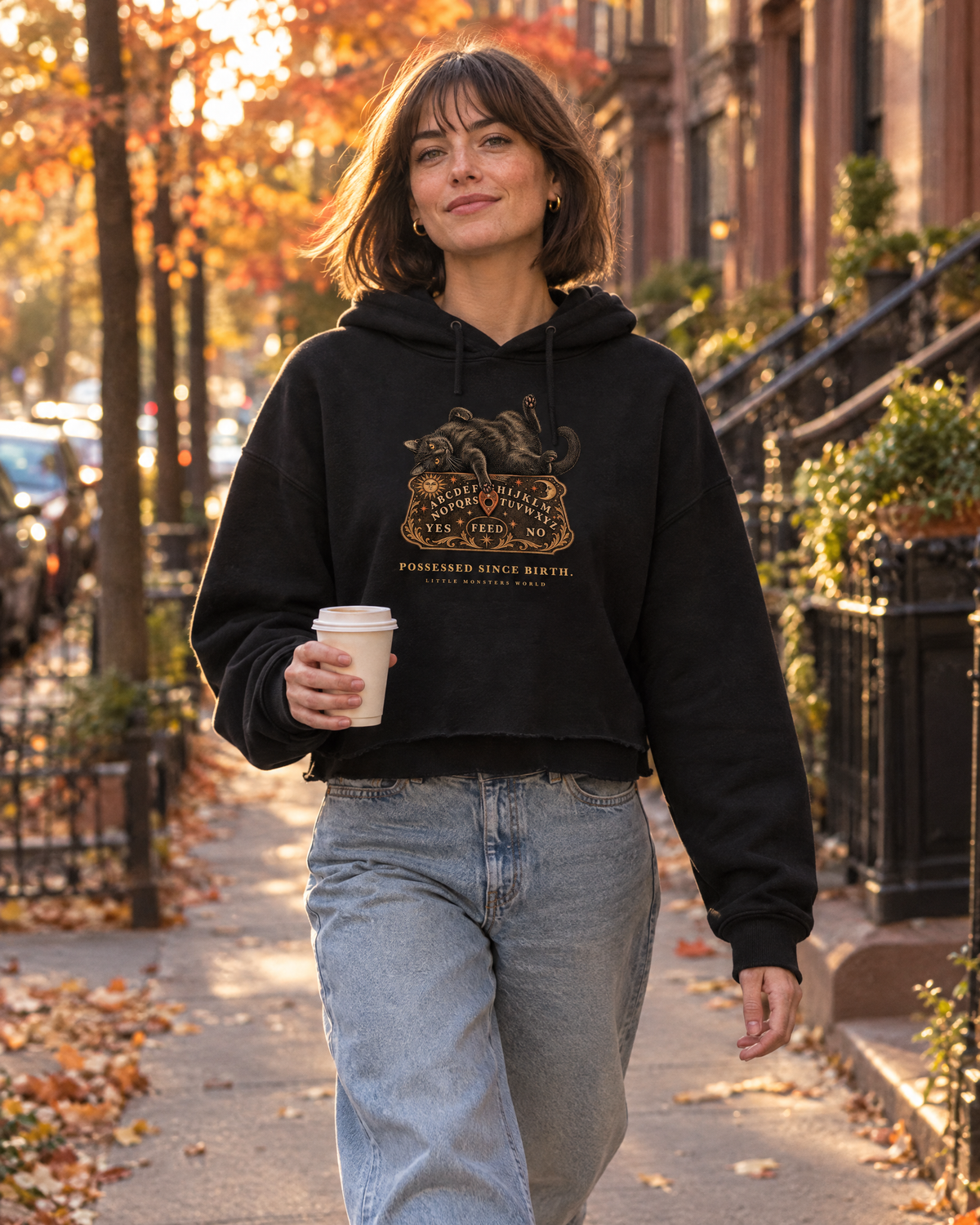 Woman walking down a brownstone street in autumn wearing the black Little Monsters Possessed Since Birth cropped hoodie