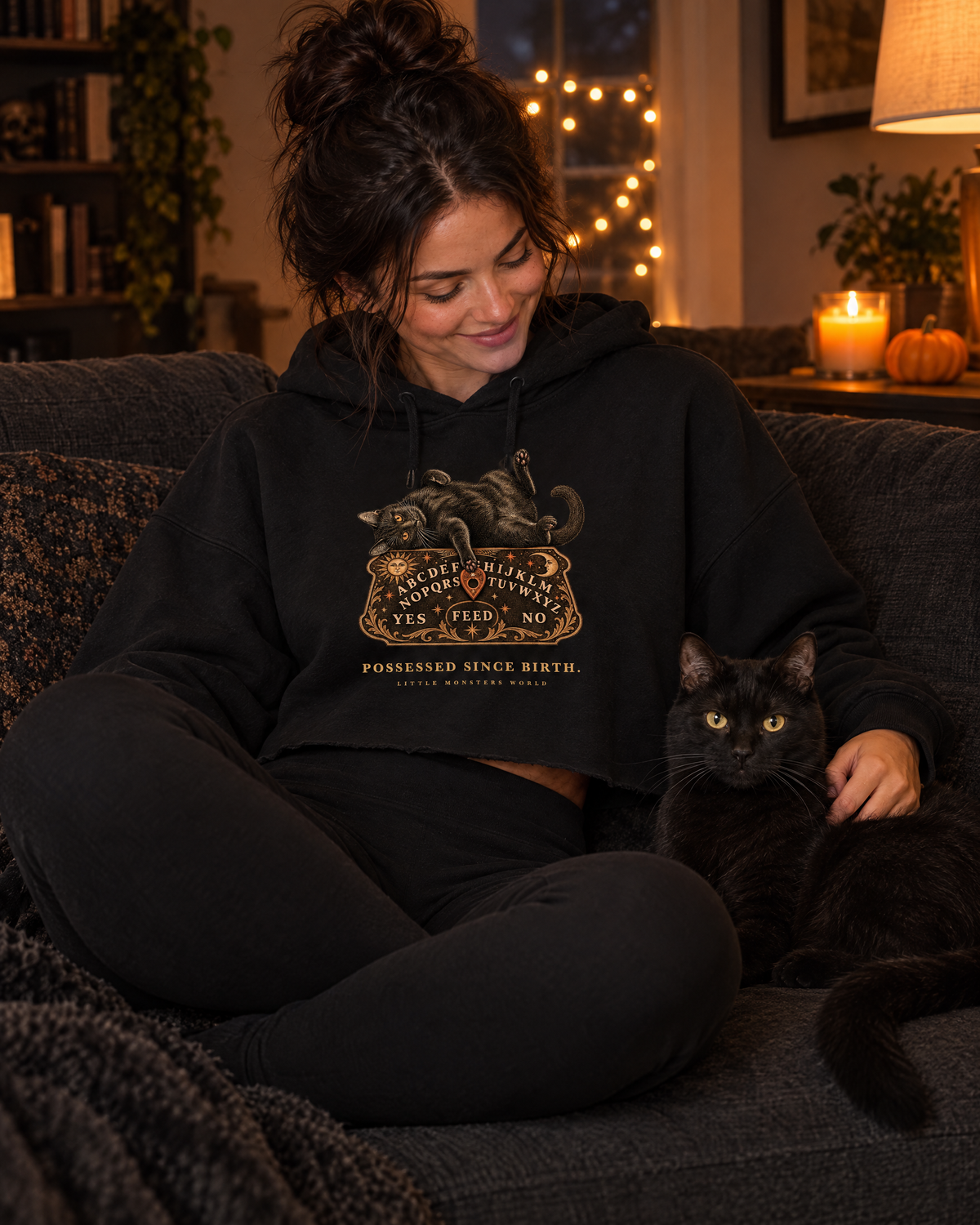 Woman on a sofa with a black cat wearing the black Little Monsters Possessed Since Birth cropped hoodie