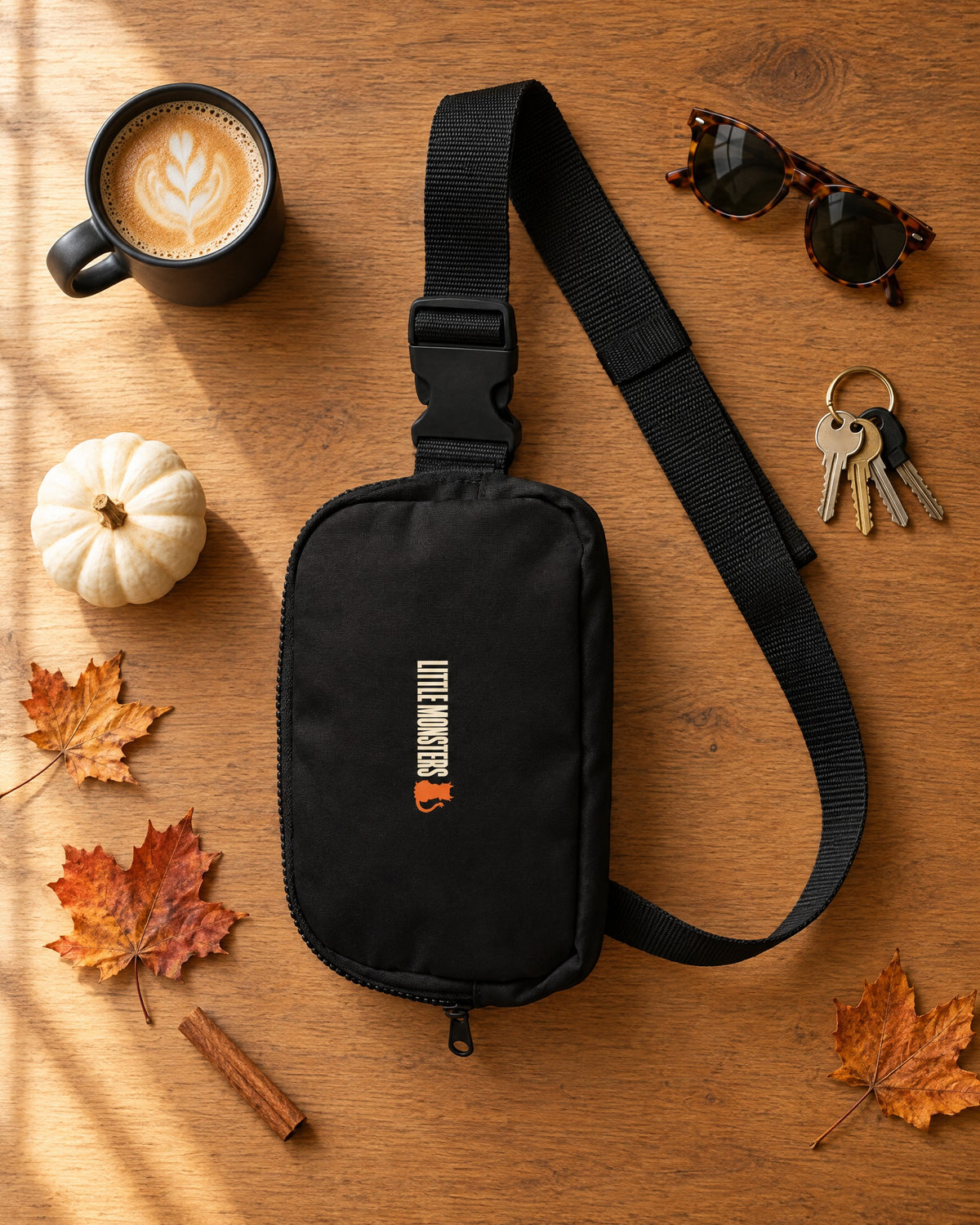 Little Monsters Logo Sling Bag in black laid flat on an oak table with a latte, sunglasses, keys, a white pumpkin and autumn leaves