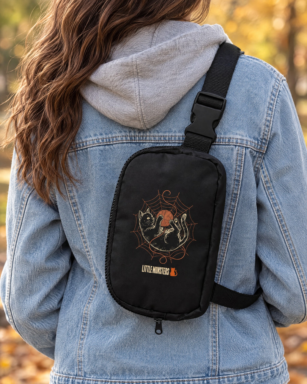 Close-up of the Yarn Web Cat Sling Bag worn over a denim jacket: a black cat in a spider web batting an orange ball of yarn, with the Little Monsters logo