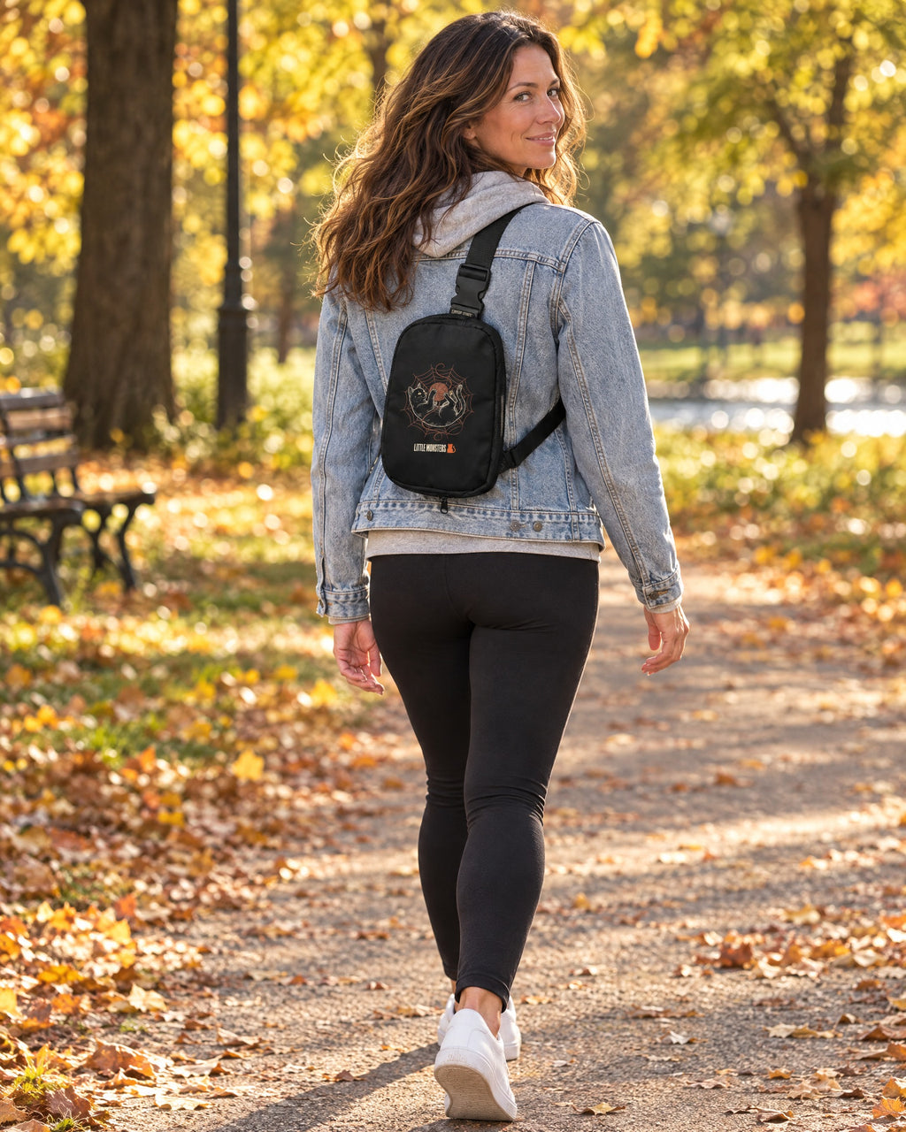 Woman in a denim jacket and grey hoodie walking through an autumn park, wearing the black Yarn Web Cat Sling Bag on her back